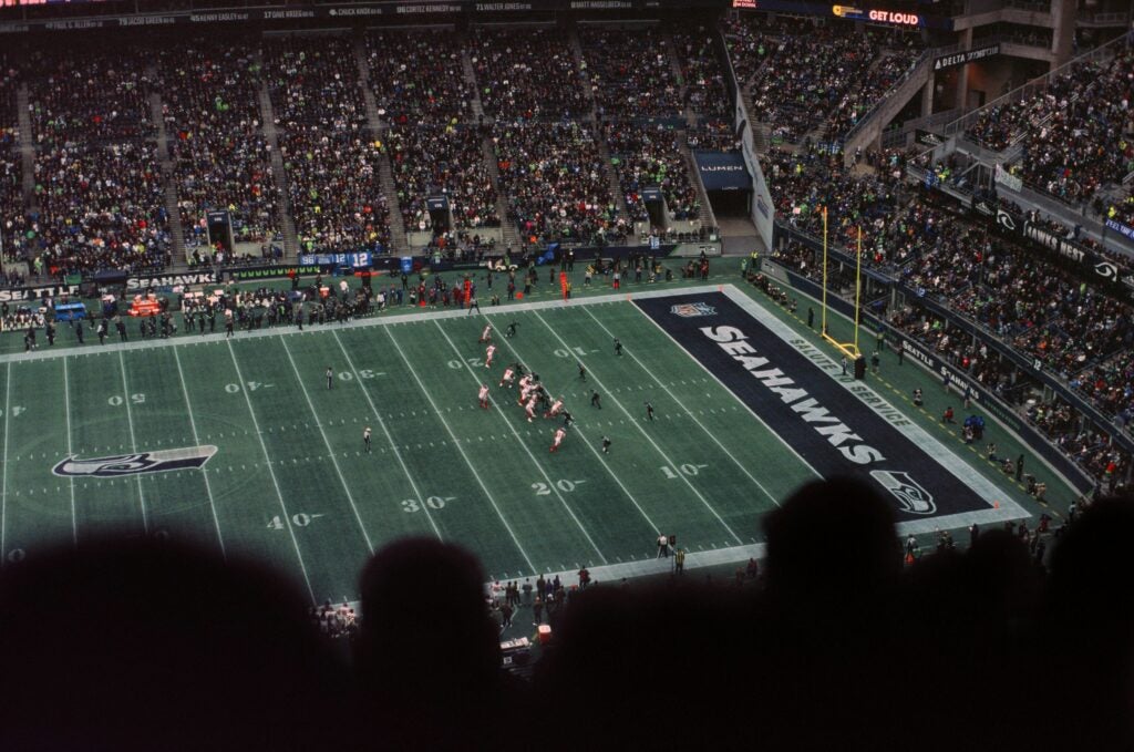 Football field in a crowded stadium with several players on the field standing at the ready. The field has the Seattle Seahawks team name and logo.