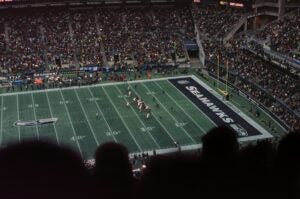 Football field in a crowded stadium with several players on the field standing at the ready. The field has the Seattle Seahawks team name and logo.