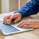 Close-up of a figure in a blue shirt, likely a man, with his hands on a laptop keyboard