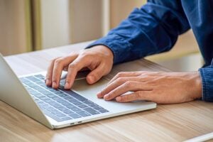 Close-up of a figure in a blue shirt, likely a man, with his hands on a laptop keyboard